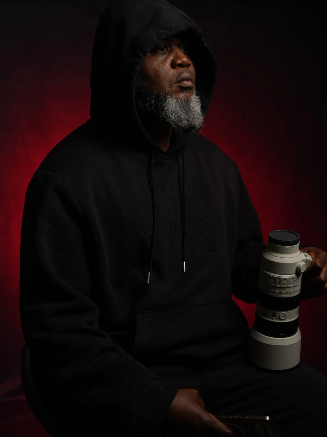 Charles Martin in a black hoodie, seated in front of a deep red backdrop, holding a camera lens.