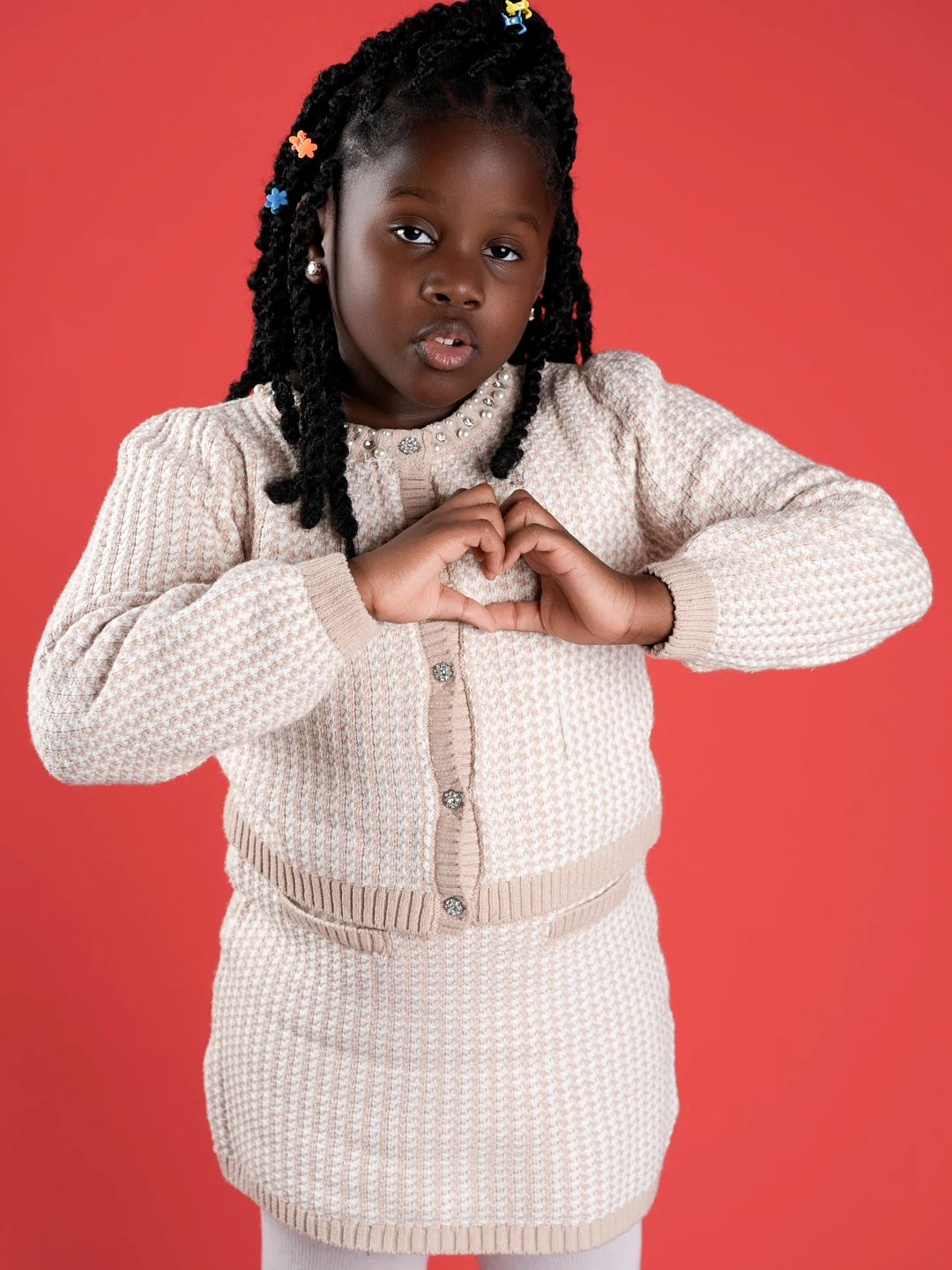 A young girl in a cream knit cardigan with pearl trim making a heart with her hands against a red backdrop.