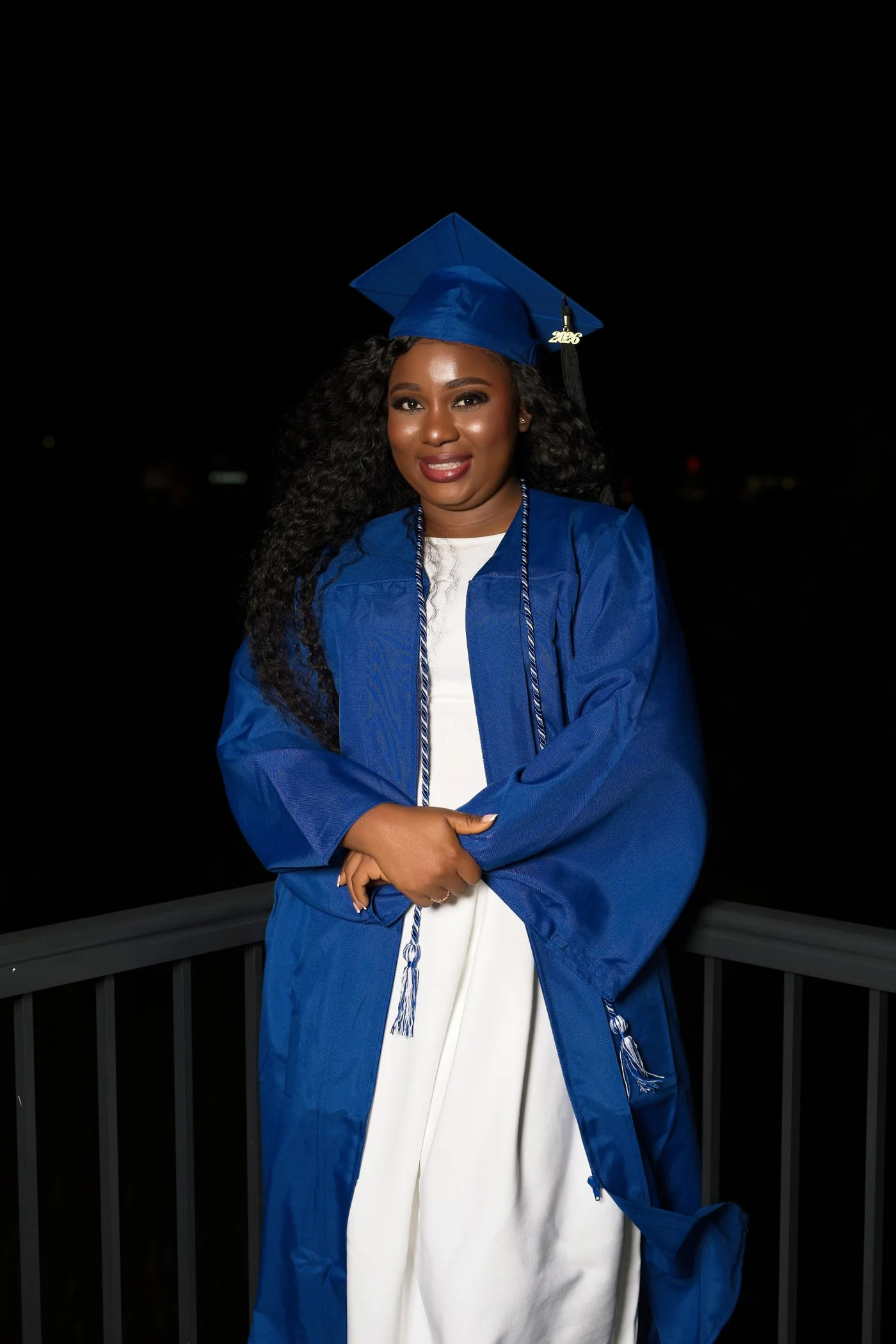 A graduate in a royal blue cap and gown over a white dress, smiling at night, lit by flash against a black sky.